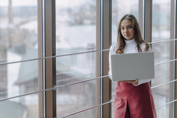 pretty young girl with laptop by the window in office with big windows. remote work concept. freelancer working alone.