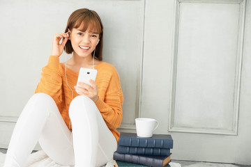 Cute asian girl wearing orange knitwear and white pants, Enjoy listening to music in the living room And a pile of books for reading,a cup of coffee,room relax,home coffee mug