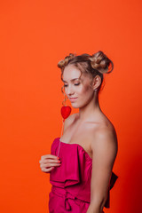 The girl in a red dress on a orange background in the studio. Blonde girl with two hair knots holding red heart on the stick, looking to the camera and smiling.