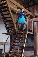 Portrait of a young girl with pink hair standing on the rusty stairs inside of collapsed building surrounded by ruins