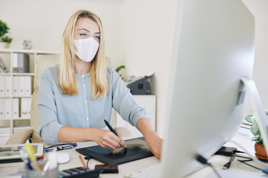 Young female graphic designer with medical mask on her face using graphics tablet while working with computer at studio or office. - Powered by Adobe
