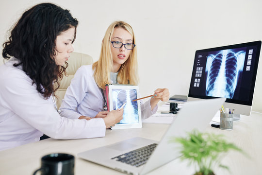 Professional young female pulmonologists pointing at lungs x-ray image on screen of tablet computer when having video conference with colleagues