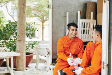 Obraz premium Cheerful young Vietnamese movers sitting in car trunk, drinking water and joking around after loading van with cardboard boxes