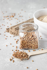 Wooden spoon with buckwheat grains and buckwheat in a glass jar, light grey concrete background. Gluten free grain for healthy diet. Selective focus.