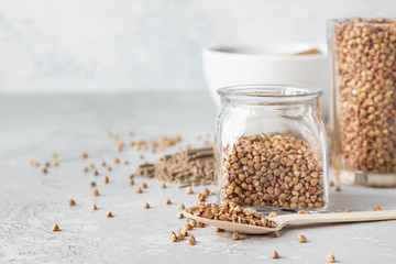 Organic uncooked buckwheat grains in glasses, buckwheat flour in a bowl and dried buckwheat soba noodles, light grey concrete background. 