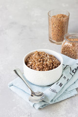 Ceramic bowl with tasty boiled buckwheat and organic uncooked buckwheat grains in glasses. Light grey concrete background. 