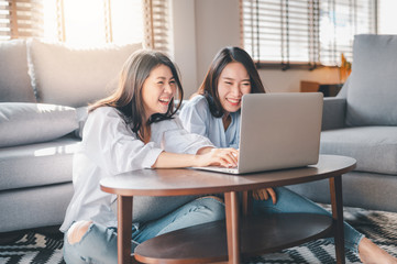 Fototapeta na wymiar Two happy Asian women best friends in casual wear laughing while working with laptop at home in living room
