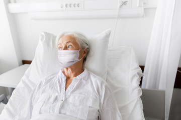 medicine, health safety and pandemic concept - senior woman patient lying in bed wearing face protective medical mask for protection from virus disease at hospital ward
