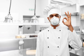Fototapeta na wymiar health protection, safety and pandemic concept - indian male chef cook wearing face protective mask or respirator showing ok gesture over restaurant kitchen background