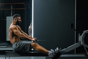 Side view of strong healthy man doing exercises with rowing machine in modern gym