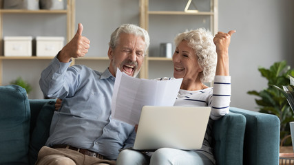 Excited mature couple reading good news in letter, using online banking service on laptop, happy...