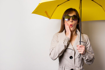 Surprised shock young woman in black sunglasses and holds a yellow umbrella on a light background. Concept of rain, bad weather