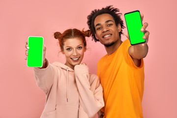 Delighted handsome black man and ginger woman points at mock up green screen of smartphone for your app, smiles joyfully, advertises new electronic device, isolated pink background. Look at cellular