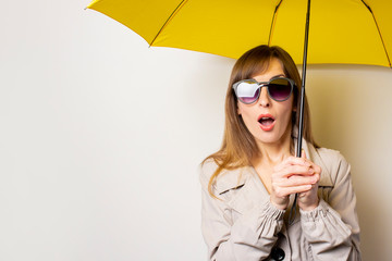 Surprised shock young woman in black sunglasses and holds a yellow umbrella on a light background. Concept of rain, bad weather