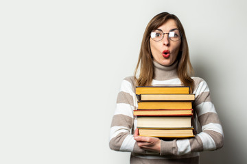 Portrait of a young friendly woman in a sweater and glasses with a surprised face holds a stack of books in her hands against an isolated light background. Emotional face. Education, exam preparation
