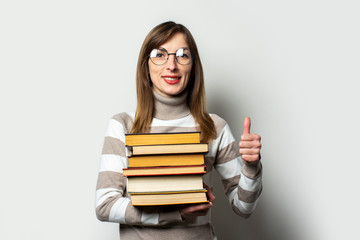 Young friendly woman in sweater and glasses holding stack of books in her hands and makes gesture Everything is fine, thumb up on light background. Emotional face. Education concept, exam preparation