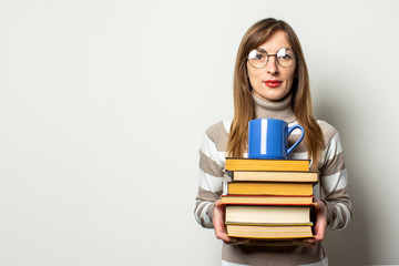 Portrait of a young friendly woman in a sweater and glasses holding a stack of books and a cup with coffee in an isolated light background. Emotional face. Education concept, exam preparation