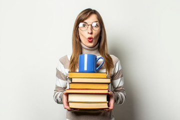 Young friendly woman in a sweater and glasses with a surprised face holds a stack of books and a cup with coffee in an isolated light background. Emotional face. Education concept, exam preparation