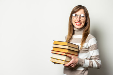 Portrait of a young friendly woman in a sweater and glasses holding a stack of books in her hands against an isolated light background. Emotional face. Education concept, exam preparation