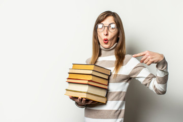 Young friendly woman in sweater and glasses with surprised face holds stack of books in her hands and points a finger at them on light background. Emotional face. Education concept, exam preparation