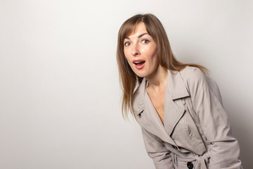 Shocked young women peeping from the side, looking at the camera. The model is dressed in a beige cloak and posing on a light background. Concept style, women's fashion