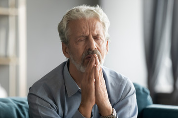 Close up sad faithful mature man with closed eyes praying with hope alone, religious upset older senior male holding hands together in prayer, begging, asking for help, saying worship