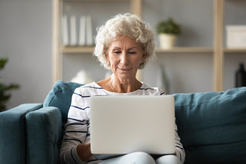 Satisfied mature woman using laptop sitting on cozy couch at home, looking at screen, chatting or shopping online, using services in internet, focused older senior female reading email