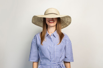 Portrait of a young friendly woman with a smile in a dress and a wide-brimmed hat on an isolated light background. The hat covers part of the face. Emotional face. Gesture of surprise, joy