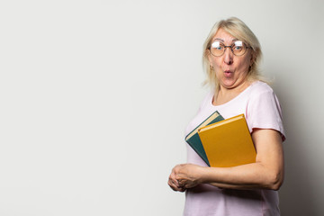 Portrait of an old friendly woman with a surprised face in a casual t-shirt and glasses holds a stack of books on an isolated light background. Emotional face. Concept book club, leisure, education