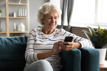 Happy smiling older woman using phone, looking at screen, sitting on cozy sofa in living room,...