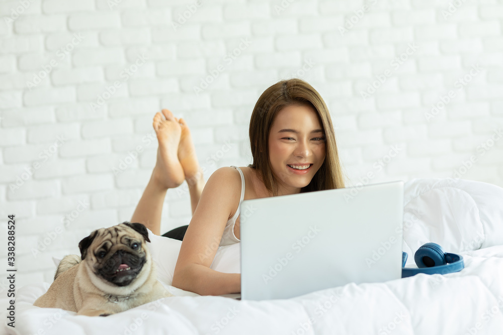 Wall mural Woman typing and working on laptop with dog Pug breed lying on bed and looking on screen feeling happiness and comfortable at home,Friendly Dog at home Concept