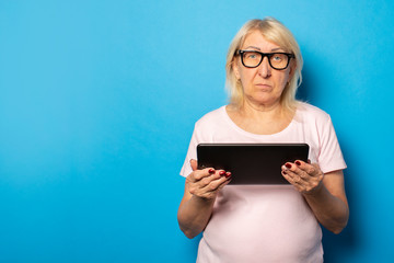 Portrait of an old friendly woman with a serious face in glasses and a casual t-shirt holding a tablet in her hands on an isolated blue background. Emotional face