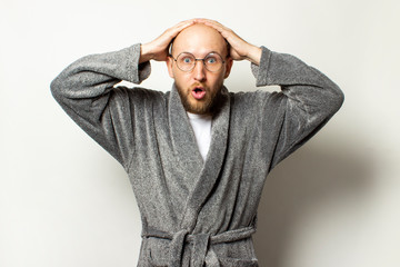 Portrait of a young bald man with a beard in a dressing gown and glasses with a surprised face on an isolated light background. Emotional face. Gesture surprise shock