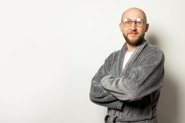 Portrait of a young bald man with a beard in a dressing gown and glasses with arms crossed on his chest on an isolated light background. Emotional face