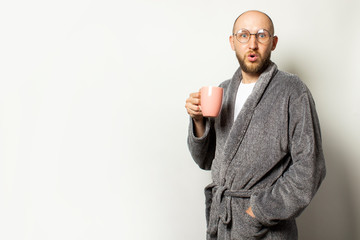 Portrait of a young bald man with a beard in a dressing gown and glasses holding a cup of hot coffee with a surprised face on a light background. Concept of morning coffee, morning, evening coffee
