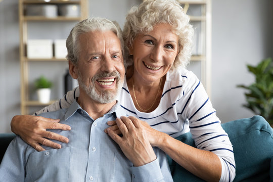 Head shot portrait loving older wife hugging husband sitting on cozy couch, happy senior mature couple with healthy toothy smiles looking at camera, posing for family photo at home