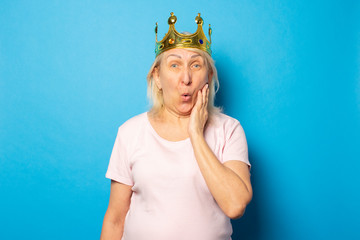 Portrait of an old friendly woman with a surprised face in a casual t-shirt with a crown on her head on an isolated blue background. Emotional face