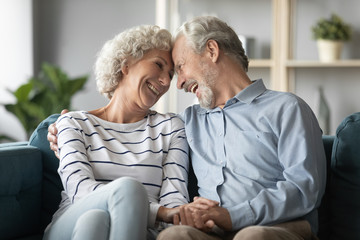 Fototapeta premium Smiling mature couple enjoying tender moment at home, older wife and husband hugging, touching foreheads, holding hands, sitting on cozy couch in living room, happy family spending time together