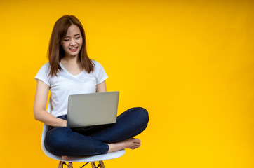 Young Asian woman teen smiling sitting on chair wearing t-shirt using laptop computer for work from home on covid-19 coronavirus crysis, studio shot on yellow background with copy space