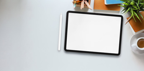 Top view image of computer tablet with white blank screen putting on white working desk and surrounded by stylus pen, potted plant, notebook, diary, coffee cup, pencils and pencil holder.