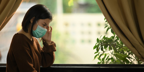 Fototapeta premium Photo of young woman wearing on medical mask feeling headache while standing at the modern living room windows. Woman keeping a quarantine herself at home. Social distancing concept.