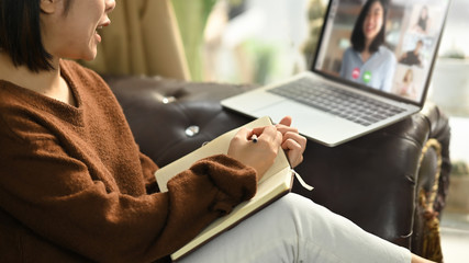 Beautiful woman taking notes while sitting in front her laptop at the leather couch in living room. Work from home/Remote working concept. Working from home through video conference concept.