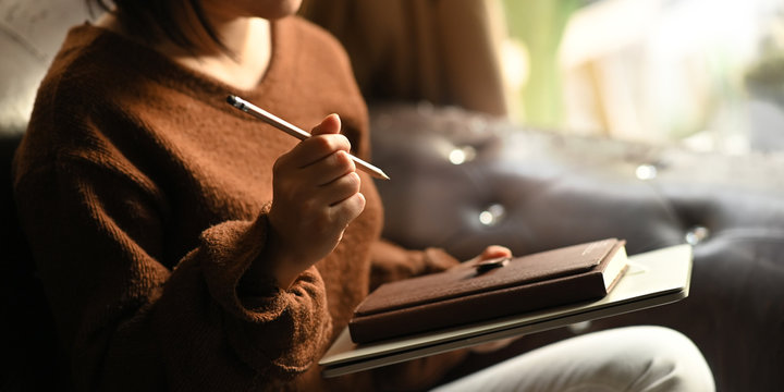 Cropped image of stylish woman holding a pencil and notebook in her hands while taking notes and sitting at the leather couch over comfortable living room as background.