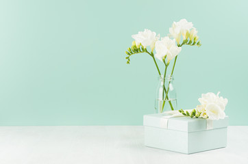 Wedding festive background with closed gift box, fragrance soft white flowers freesia in glass vase in green mint menthe interior on white wood board.