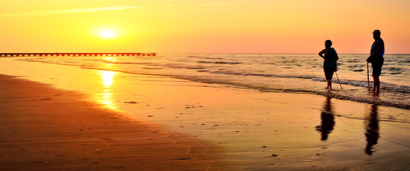 Fototapeta premium Banner, web page or cover template. Standard size. Retired people walking on the beautiful morning beach during sunrise. Copy space and panoramic ratio
