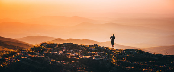 Banner, web page or cover template of Alone man standing on the top of the hill on the mountains meadow with beautiful colorful sunset over landscape. Copy space and panoramic ratio