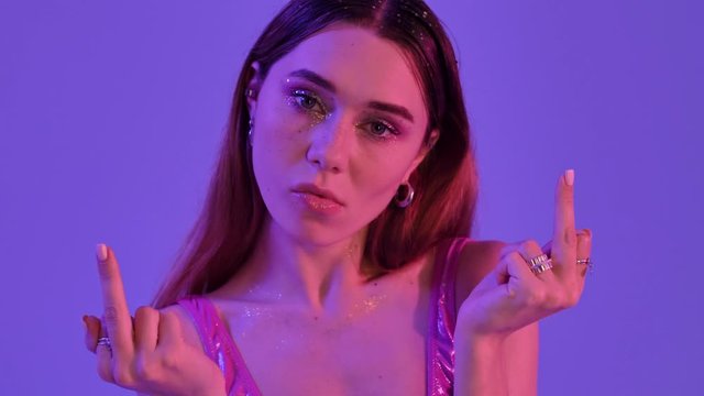 A serious confident woman with fashion glitter makeup is showing a middle finger gesture with both hands isolated over violet neon background