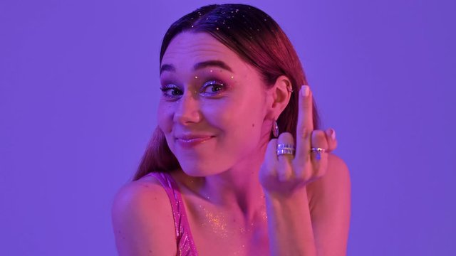 A confident woman with fashion glitter makeup is showing a middle finger gesture isolated over violet neon background