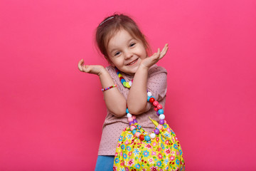 Studio shot of cute charming little lady wearing casual outfit and different multicolored jewelry, keeping hands near face, looking smiling directly at camera, having funny facial expression. - obrazy, fototapety, plakaty