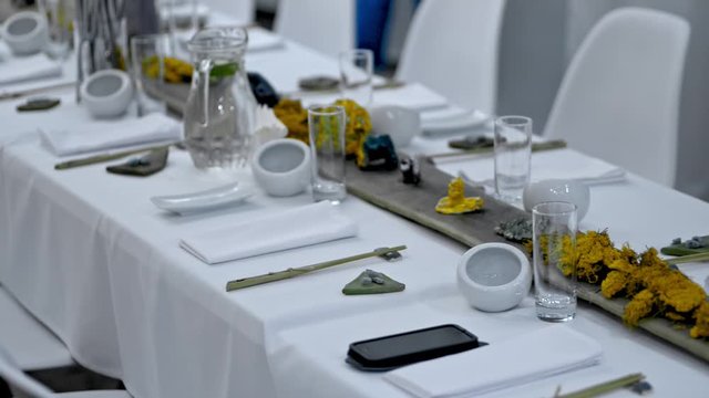Beautiful view of festive table with arrangement for event in luxury restaurant irrl. Crockery and glasses, decorative items and drinks are on white tablecloth of long desk with chairs. Modern serving